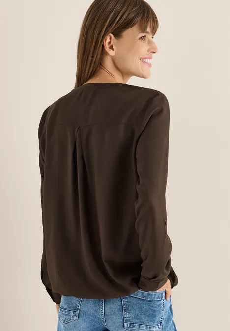 Frau mit brauner Bluse und Jeans, lächelt und schaut über die Schulter.